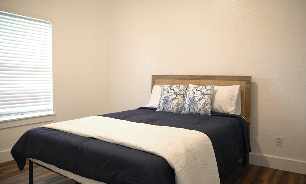 A simple, clean bedroom featuring a wooden bed with dark blue bedding and white accent pillows. Natural light filters through a window with blinds, creating a calm and mini