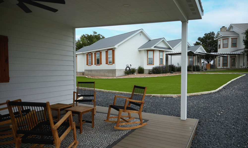 A view from a covered porch looking out at a set of white residential buildings with green lawns and gravel paths. Wooden rocking chairs sit on the porch, conveying a peaceful, community-oriented recovery setting.
