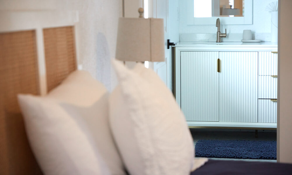 A softly lit bedroom scene with a focus on a bed and white pillows in the foreground, leading into a clean, modern bathroom with a white vanity and gold hardware. The composition highlights comfort and tranquility in a residential treatment environment.