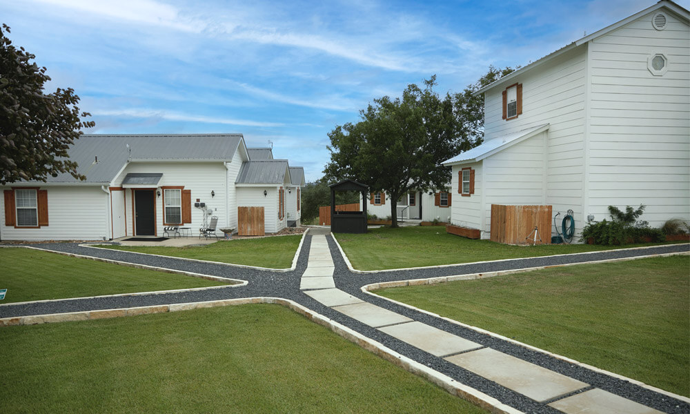 A peaceful, well-maintained property with several white buildings featuring wooden shutters and metal roofs, connected by clean stone walkways. The open, green landscape and tranquil setting evoke a sense of calm and renewal — an ideal environment for rehabilitation and recovery.
