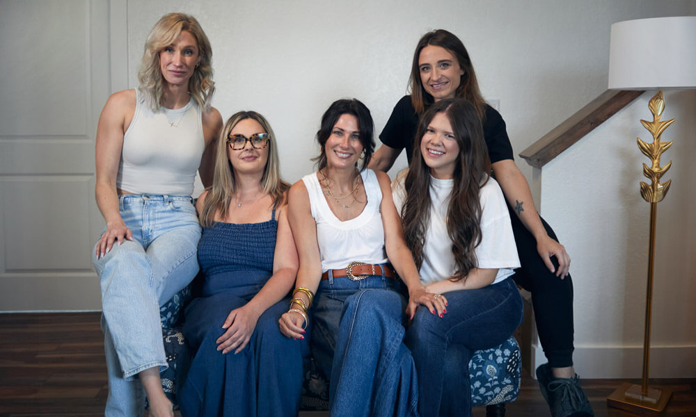 A group of five women sitting together indoors, smiling warmly and appearing relaxed. They are dressed casually in jeans and light tops, creating a welcoming, supportive, and friendly atmosphere that reflects a women-focused recovery or wellness environment.