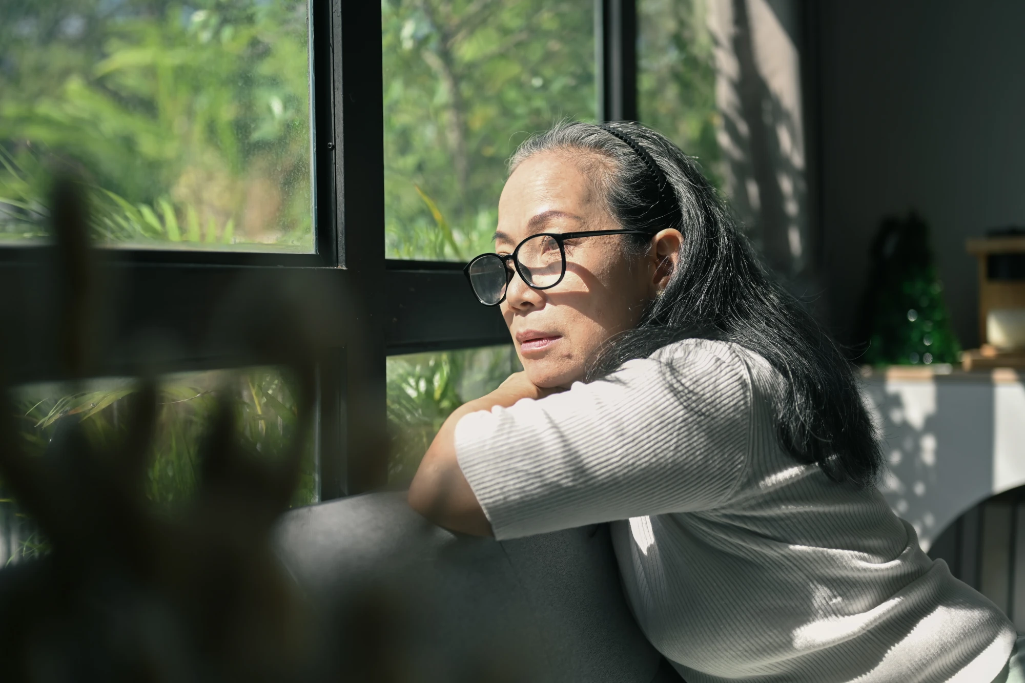 An older woman sits by a window with a thoughtful expression.